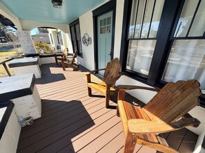 Una terraza de madera con sillas Adirondack, una mesa, una puerta y ventanas con persianas.