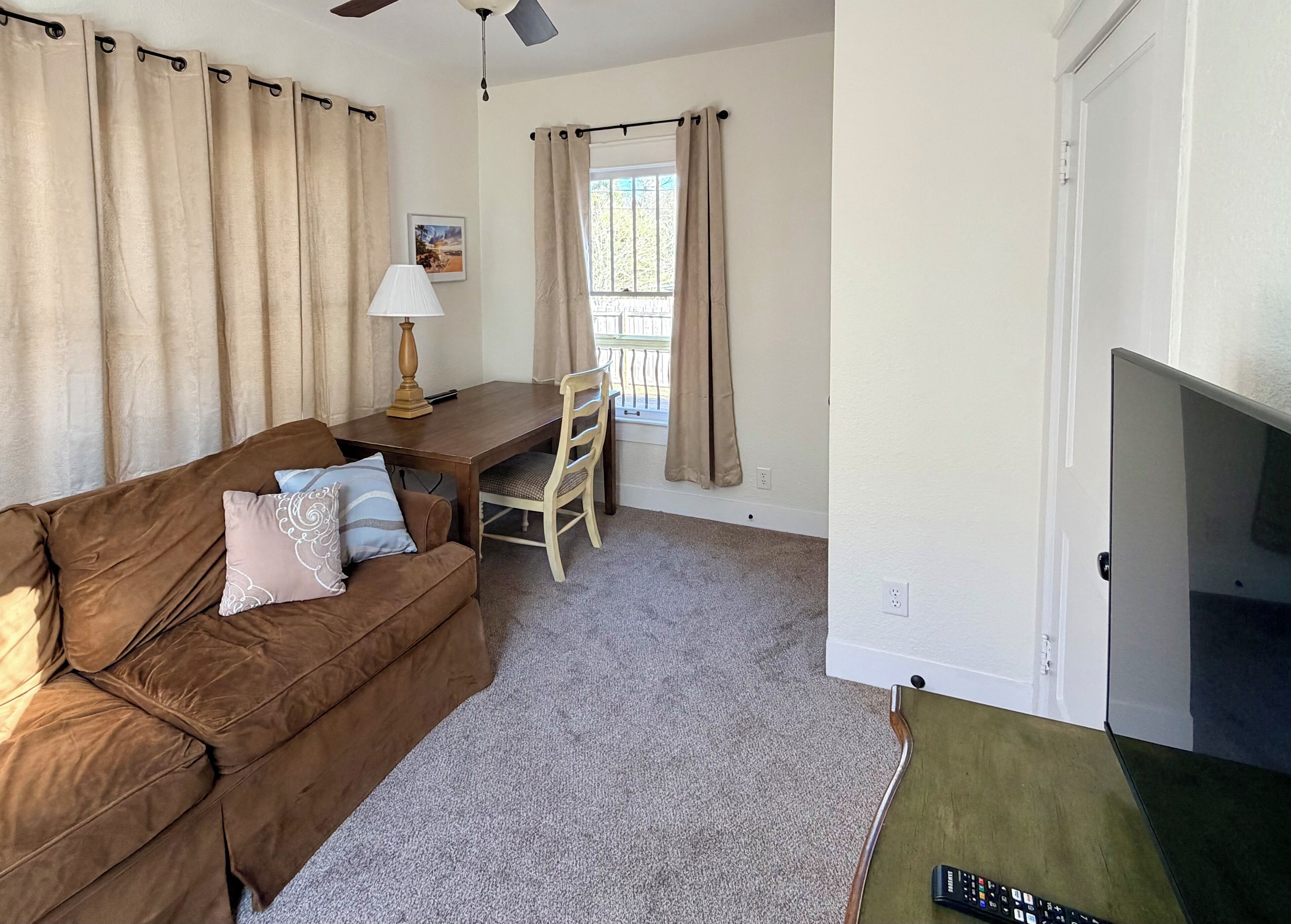 A living room with a brown sofa, a small table with a lamp, a TV, and beige curtains.