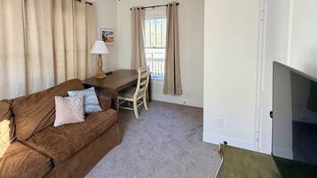 A living room with a brown sofa, a small table with a lamp, a TV, and beige curtains.
