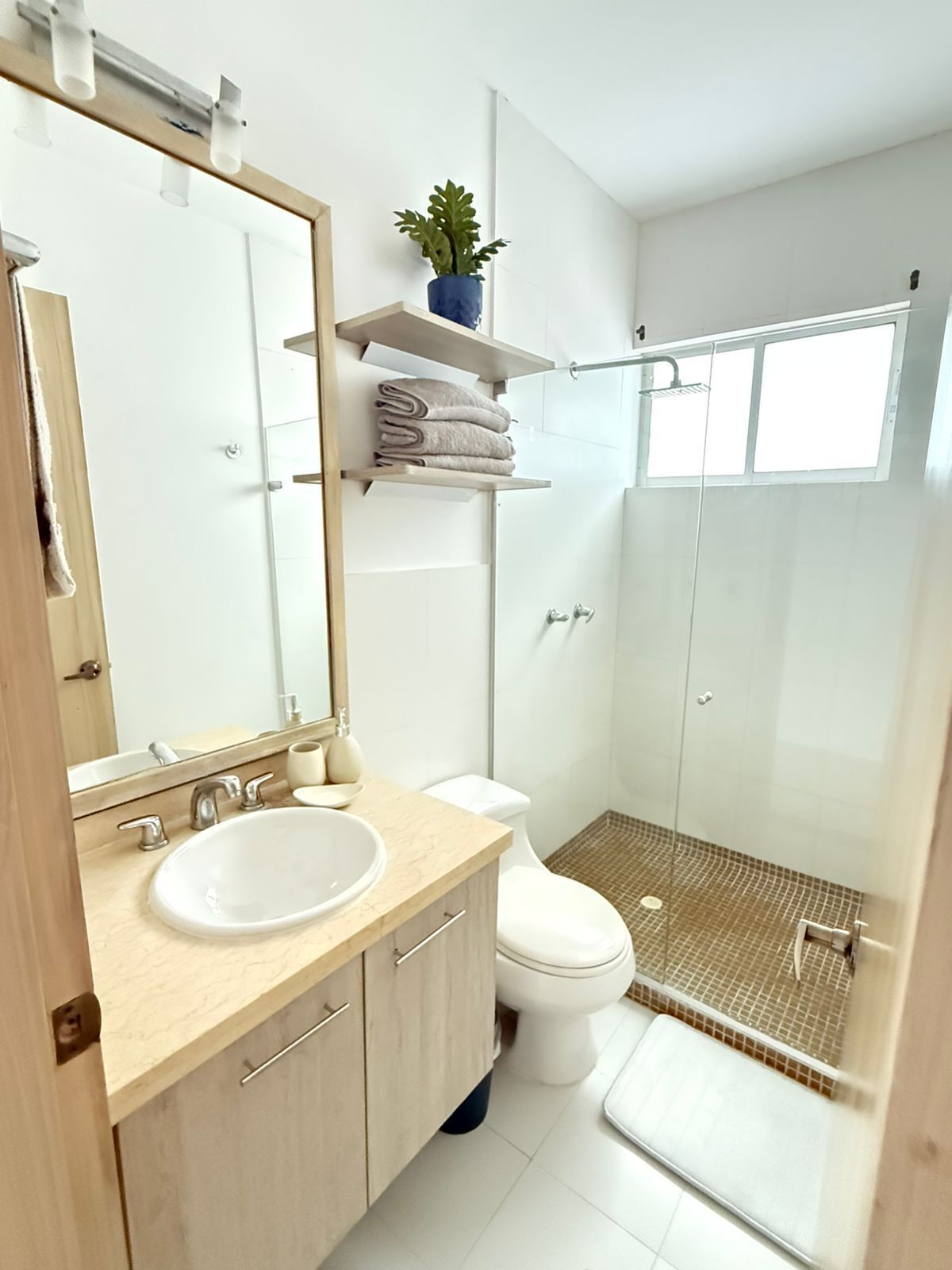 Ein modernes Badezimmer mit Waschbecken, Spiegel und Toilette. Es gibt eine Dusche mit Glastür sowie eine kleine Topfpflanze auf einem Regal.
