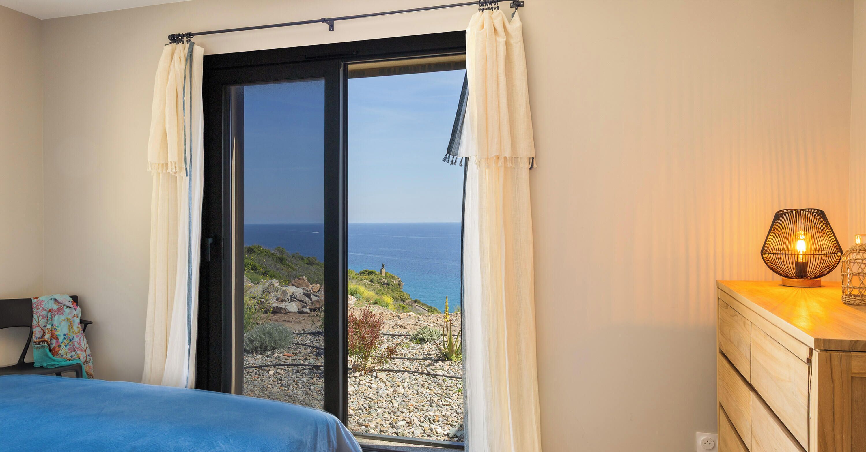 Un dormitorio con cama, cómoda, lámpara y vista al mar a través de una puerta corrediza.