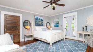 Une chambre à coucher comprenant un lit, un ventilateur de plafond, une table de chevet, une chaise, une table et des œuvres d’art accrochées aux murs.