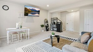A modern living room with a flat-screen TV, a dining area with a white table and chairs, a sofa, and a kitchen area with a microwave and shelves.