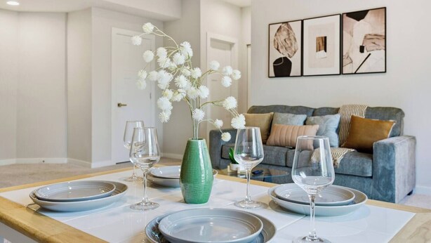 A dining area with a table set for four, a green vase with white flowers, and a grey sofa in the background.