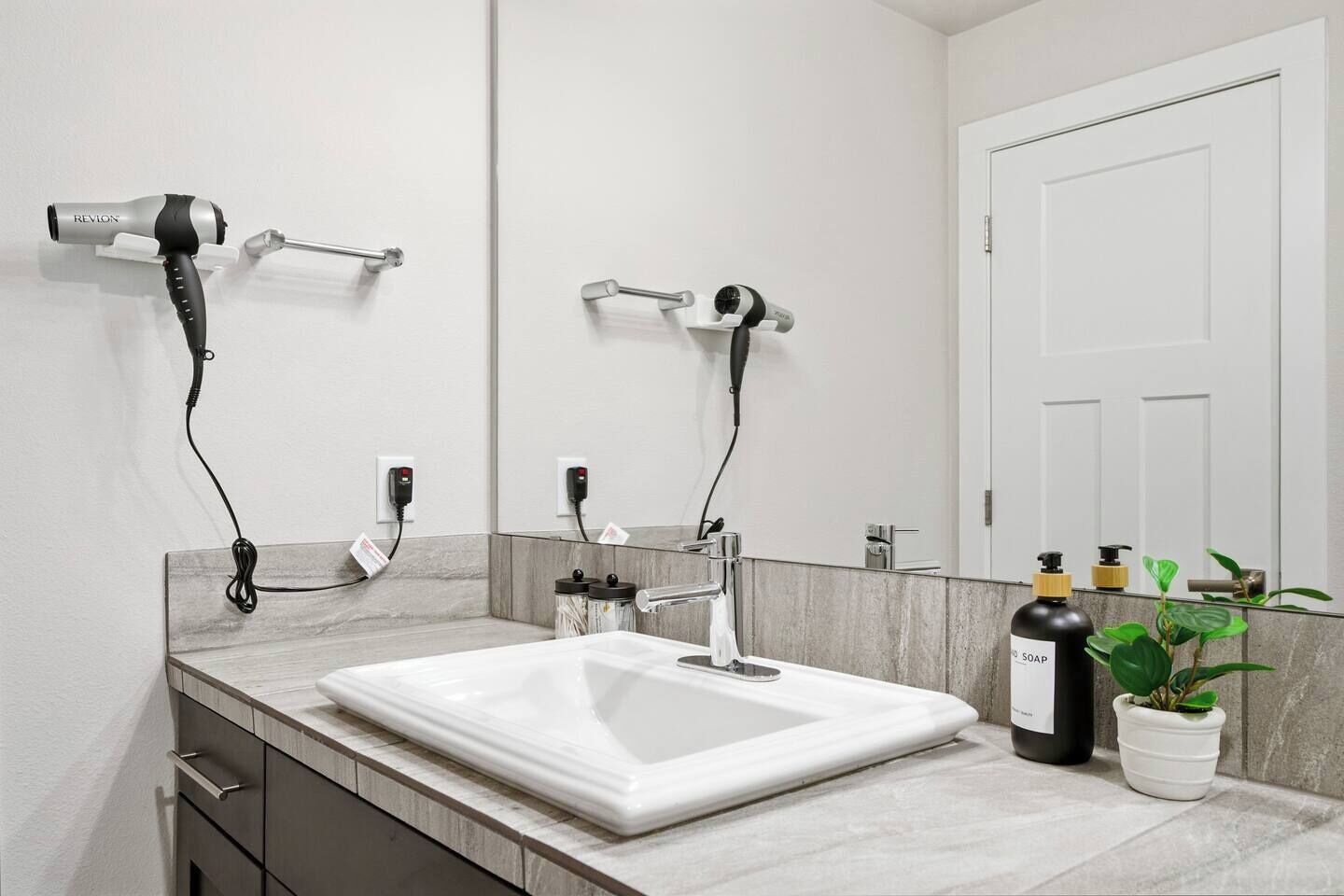 A bathroom with a large mirror, two hair dryers mounted on the wall, a white sink, and a potted plant.