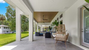 Überdachte Terrasse im Freien mit Deckenventilator, Korbmöbeln und Blick auf ein Haus und Grünflächen.