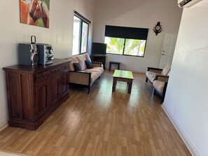 A living room with a wooden cabinet, a beige sofa, a small wooden table, and a window with a view of greenery. - The Ningaloo breeze 2 (Exmouth)