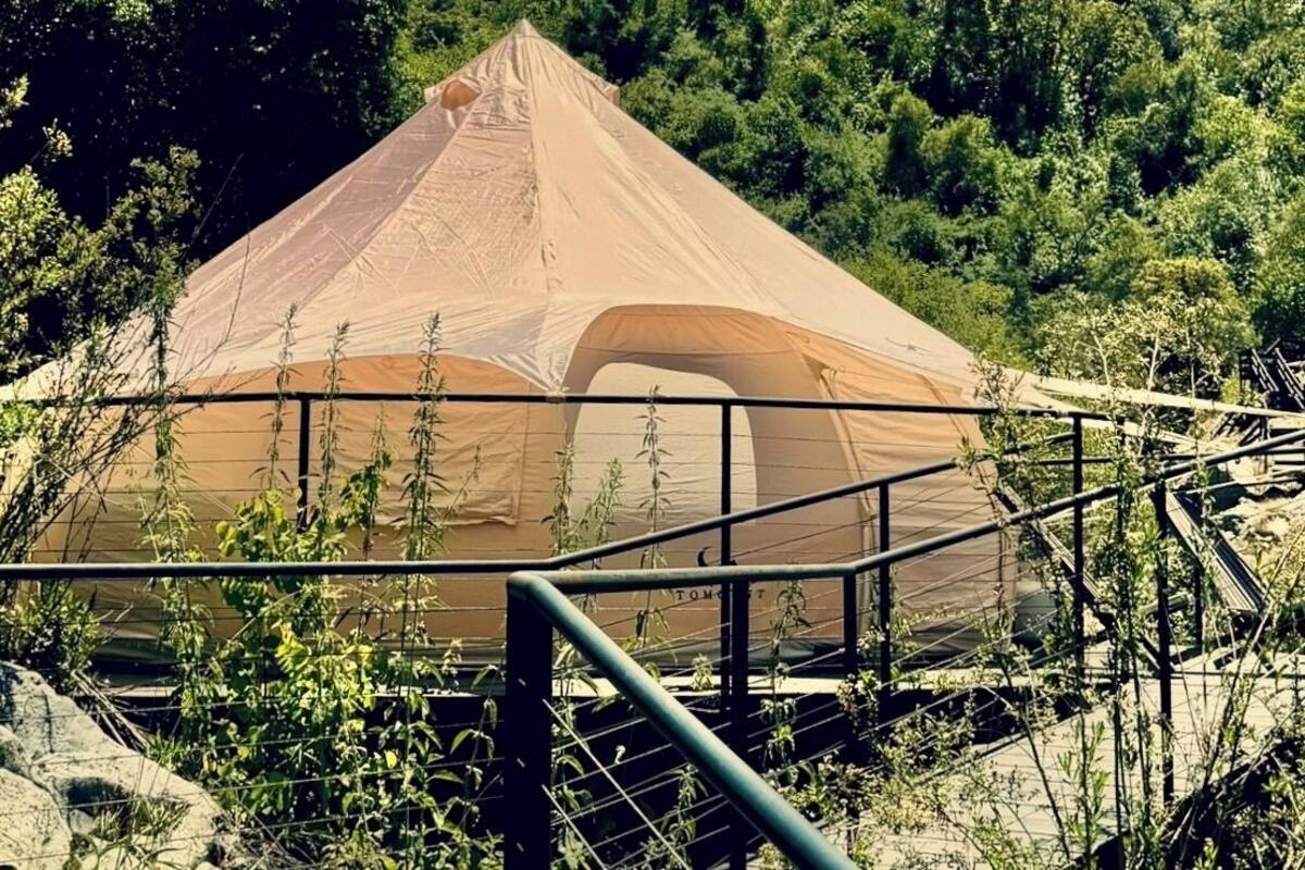 A large tent with a peaked roof, surrounded by a metal railing, set in a natural, wooded area.