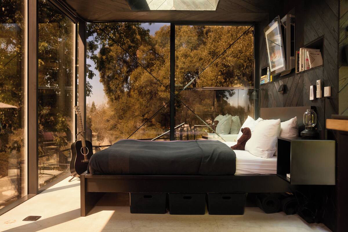 A modern bedroom with a bed, a guitar, and a view of trees through large windows.