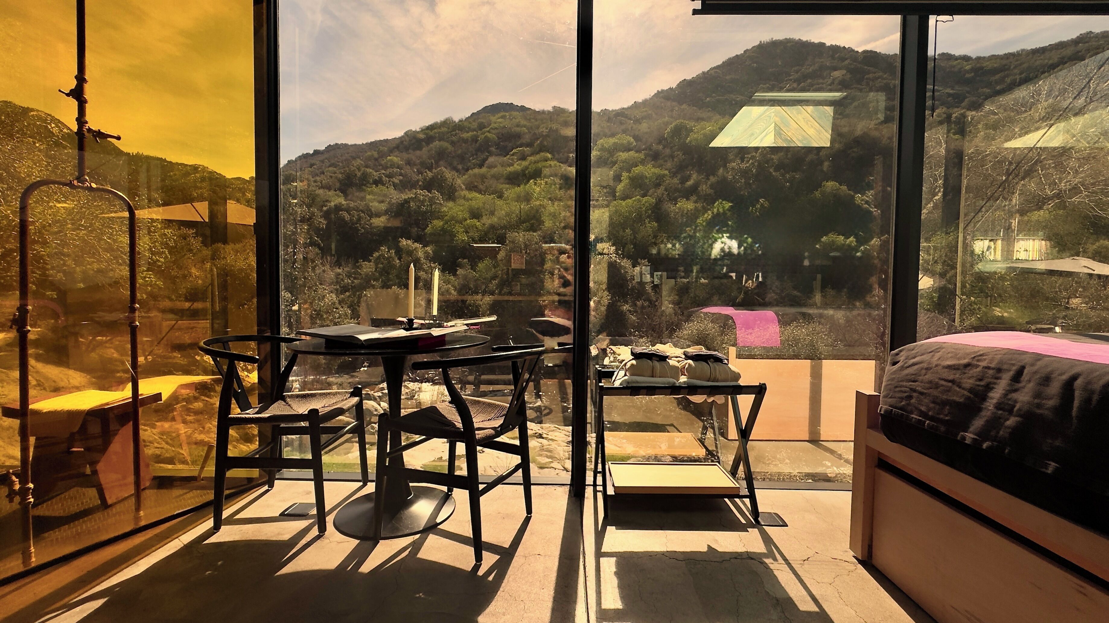 A room with a large window offering a view of a mountainous landscape. Inside, there is a bed, a table with chairs, and a small table with a candle.