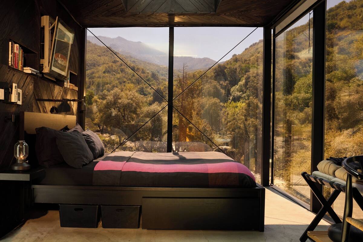 A bedroom with a large window, a bed, a nightstand, and a bookshelf.