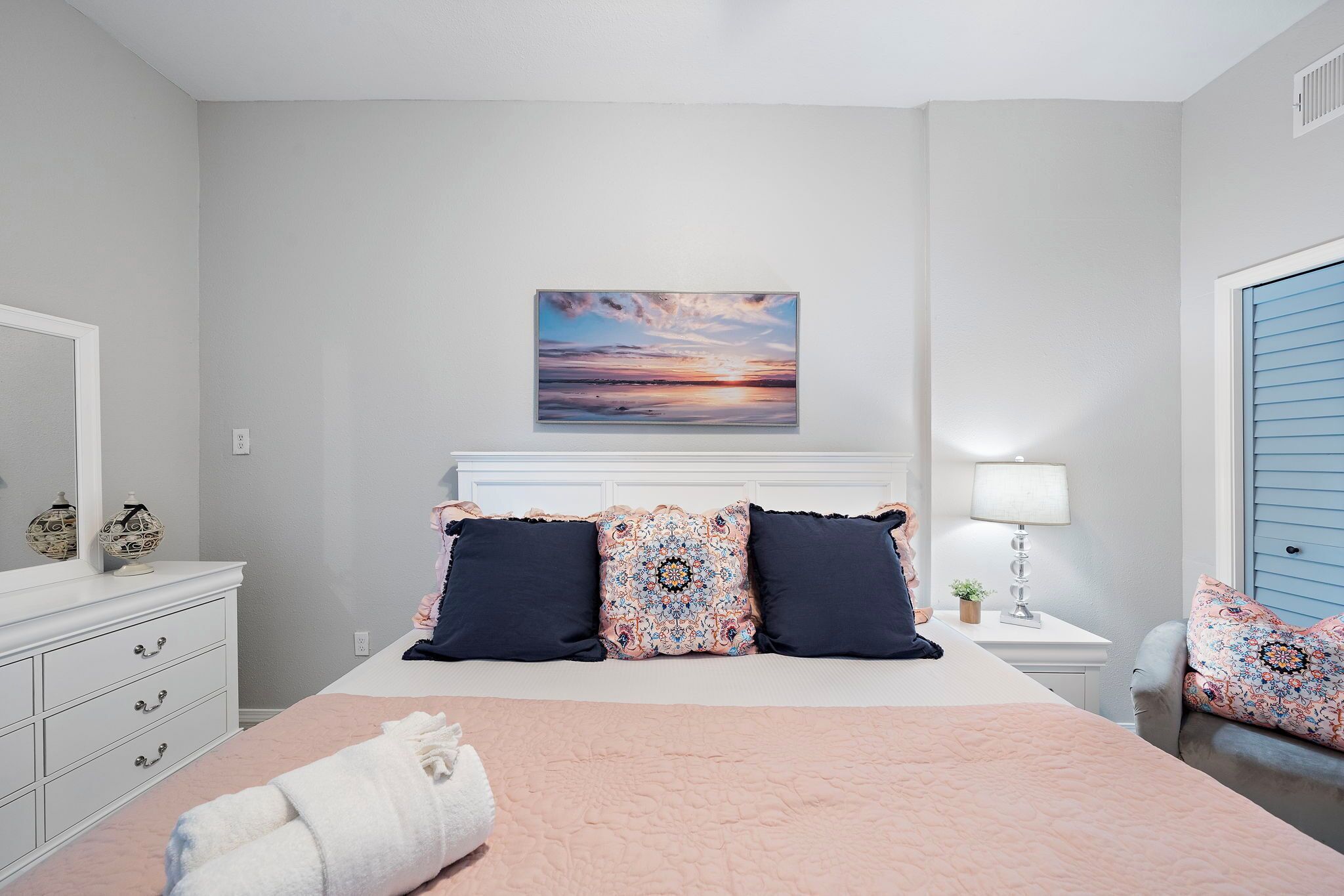 A bedroom with a bed, a nightstand, a lamp, a dresser, and a window with blinds.