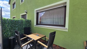 Une terrasse avec une table et des chaises, une fenêtre avec des stores et un mur végétal.