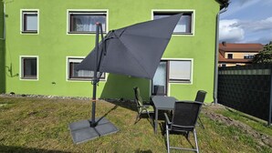 Un patio con una mesa y sillas, un paraguas grande, y un edificio verde con ventanas al fondo.