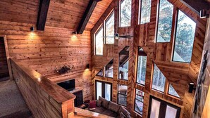 L’intérieur d’une cabane en rondins, avec une chambre à coucher située en mezzanine, de grandes fenêtres et une cheminée.