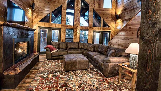 L’intérieur d’une cabane en rondins, avec un grand canapé d’angle en forme de L, une cheminée et un tapis à motifs.