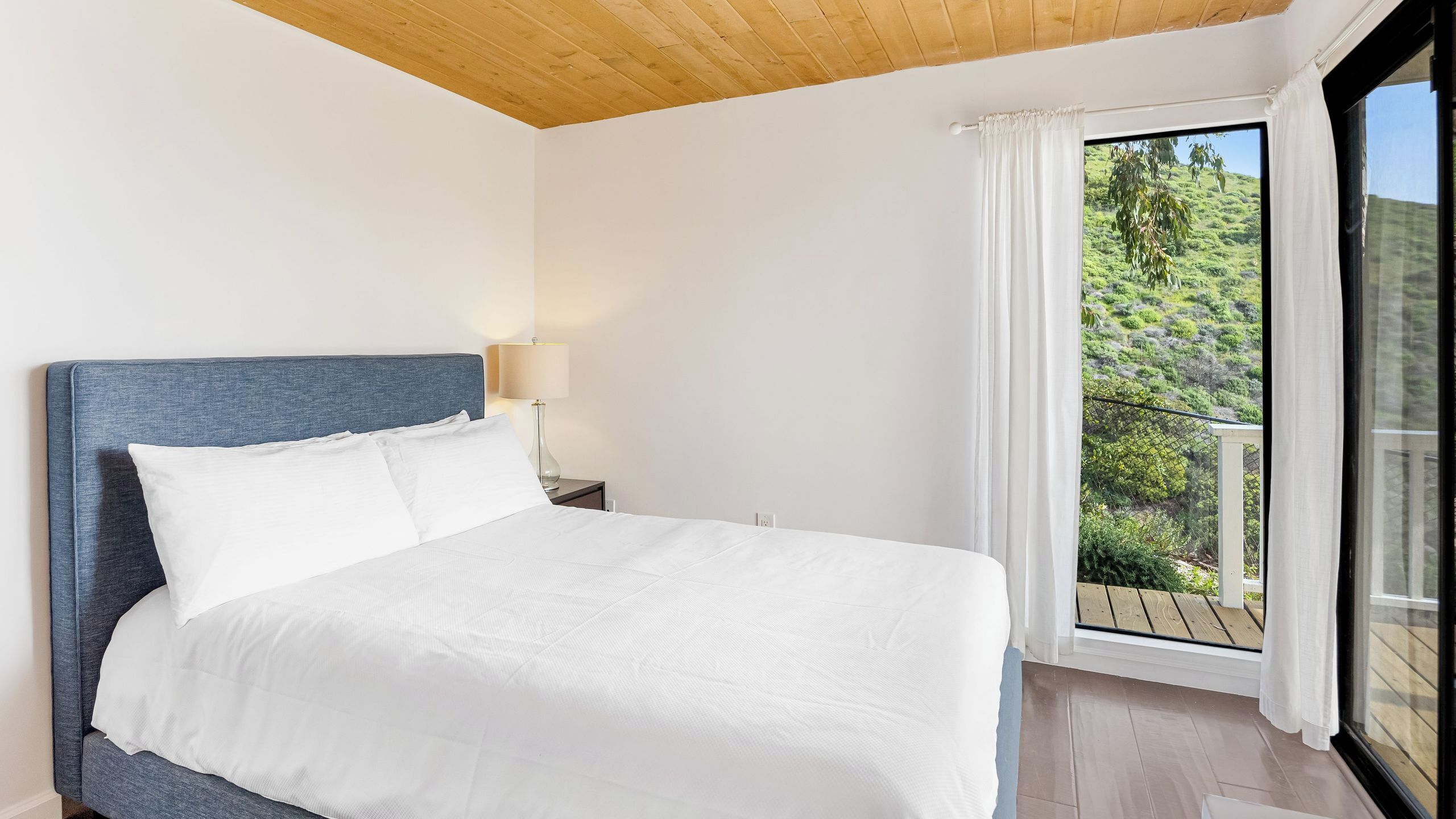 Ein Bett mit weißer Bettwäsche, eine Holzbalkendecke, ein Nachttisch mit Lampe und ein Fenster mit Blick auf Grün.