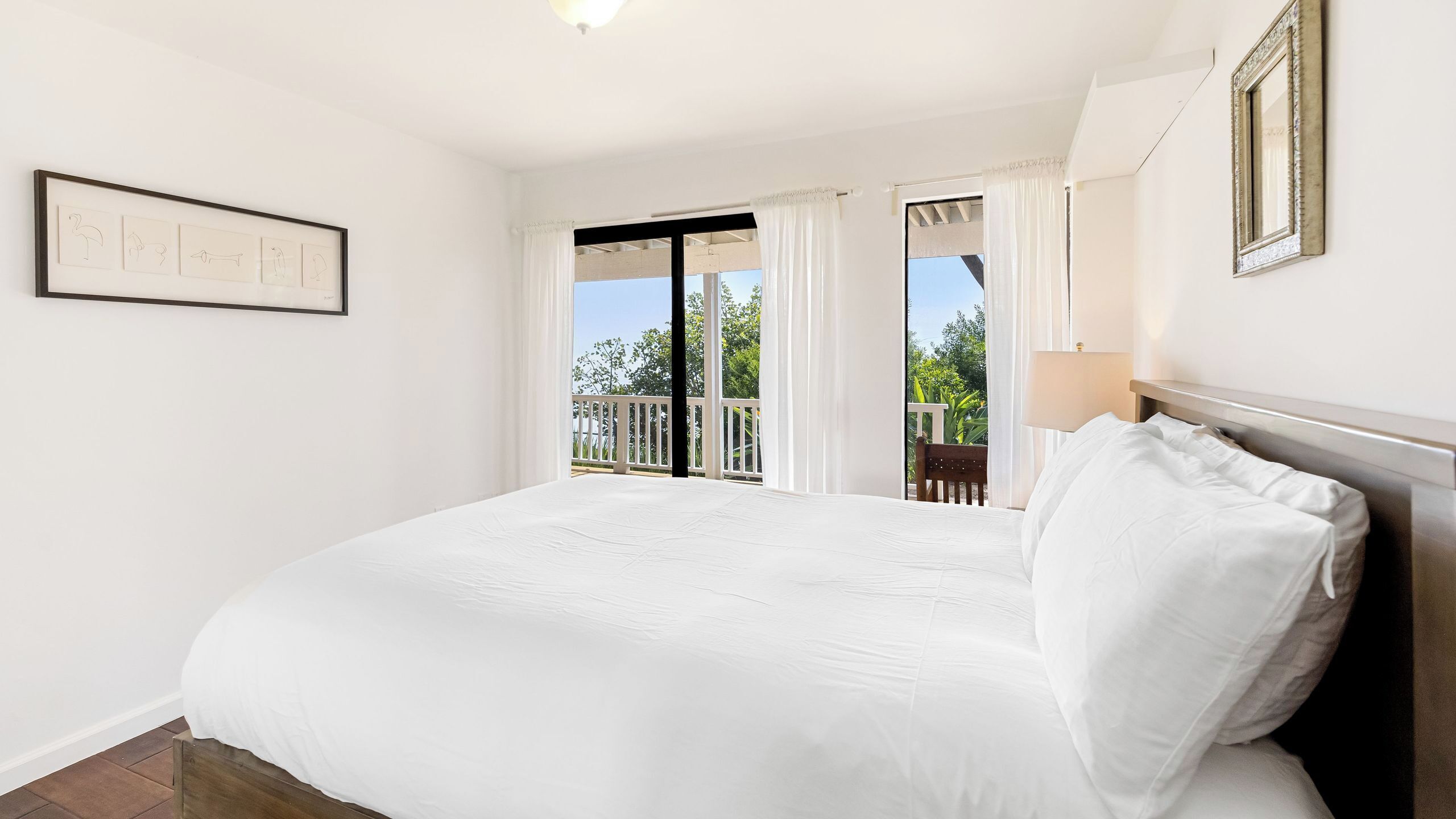 A bedroom with a large bed, a framed picture on the wall, and a view of a balcony with a railing.