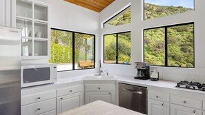 A modern kitchen with white cabinetry, a built-in microwave, a coffee maker, and large windows offering a view of greenery. - Pacific View Malibu by Avantstay Bluff-top Ocean Views + Private Pool & Hot Tub (Malibu)