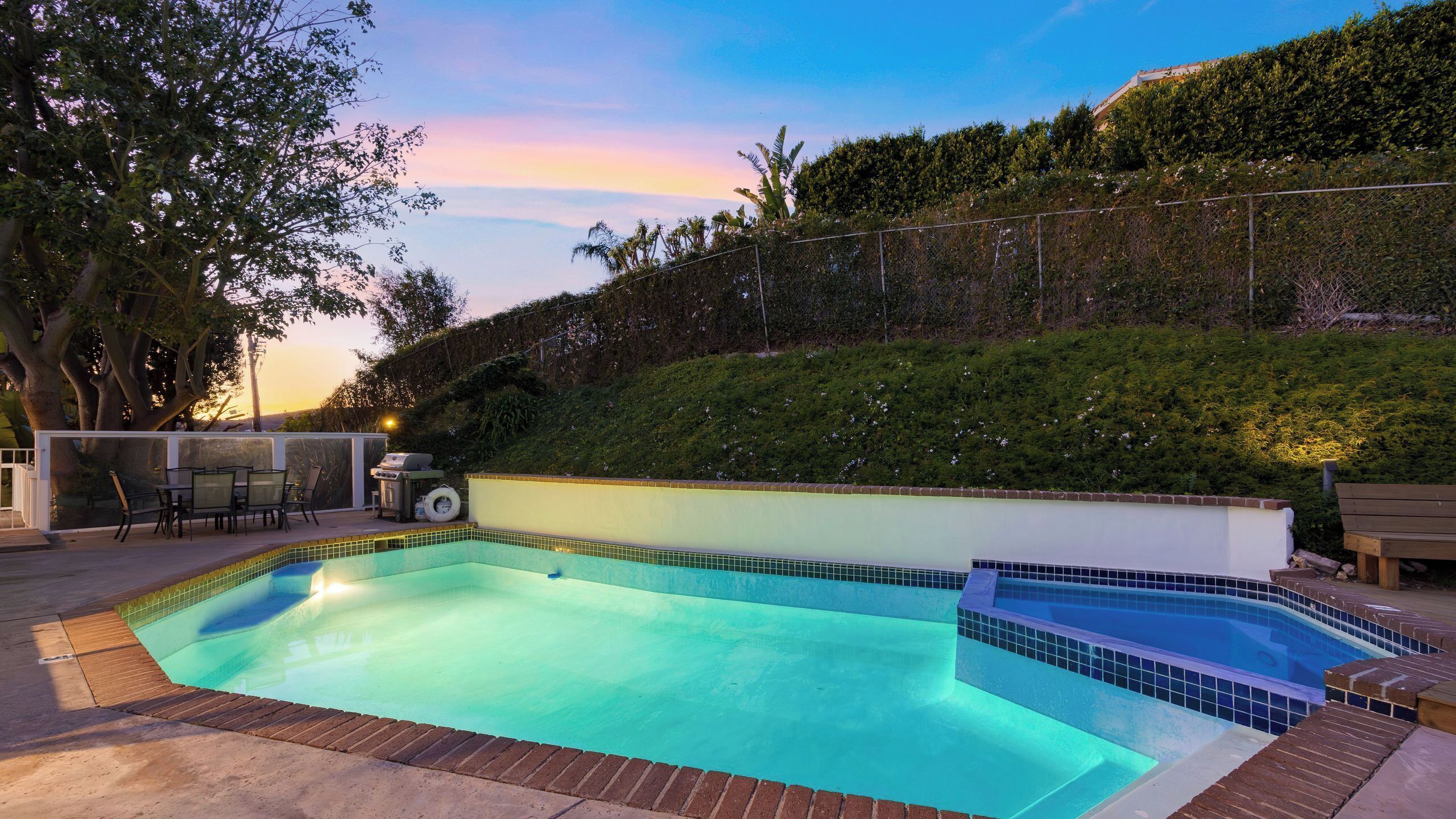 A swimming pool with a tiled edge, surrounded by a patio area with chairs and a grill.