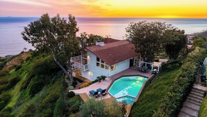 Una casa de dos pisos con piscina, rodeada de abundante vegetación y vistas al océano.