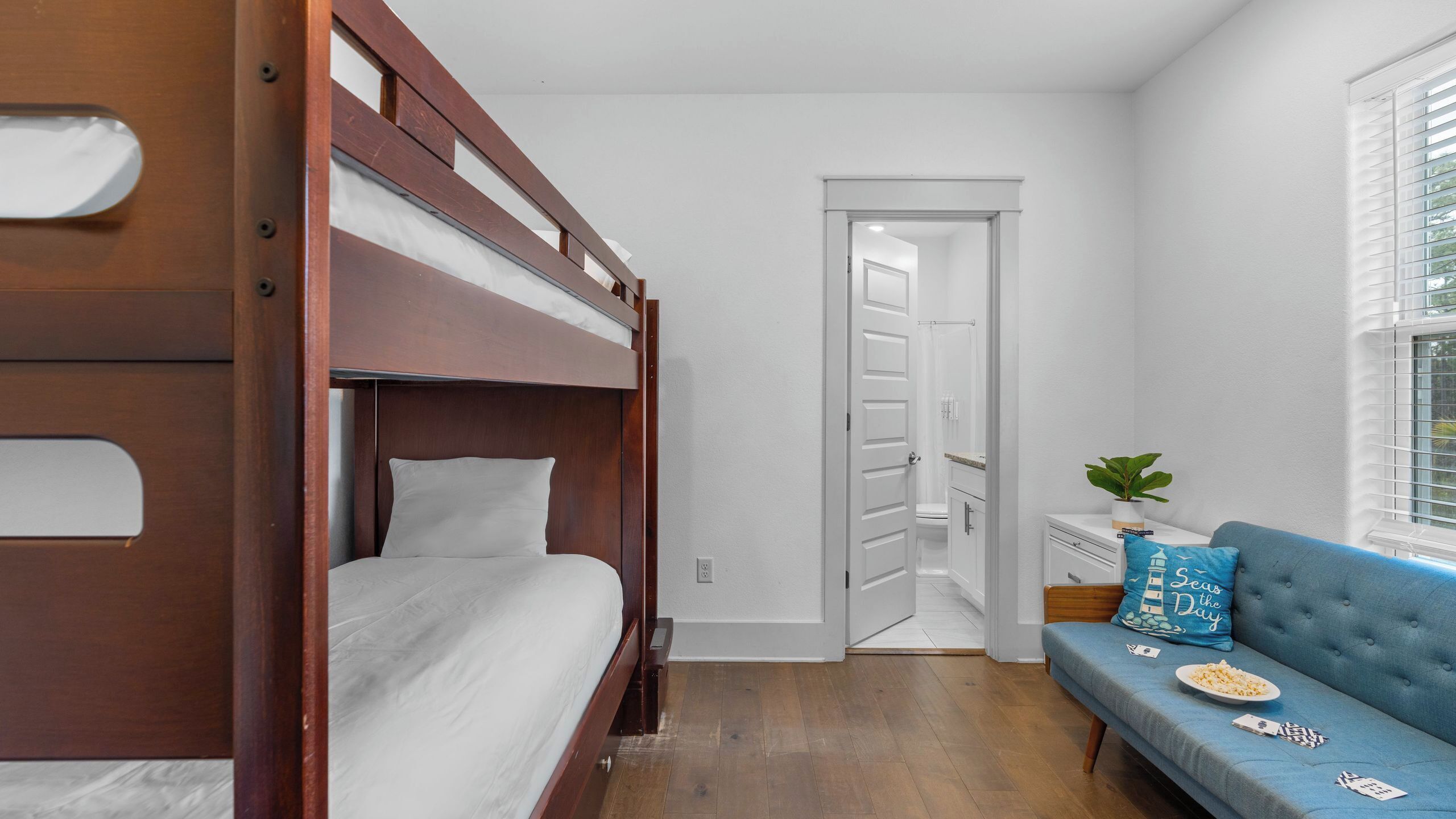 A bedroom with a bunk bed, a sofa, a window with blinds, and a door leading to a bathroom.