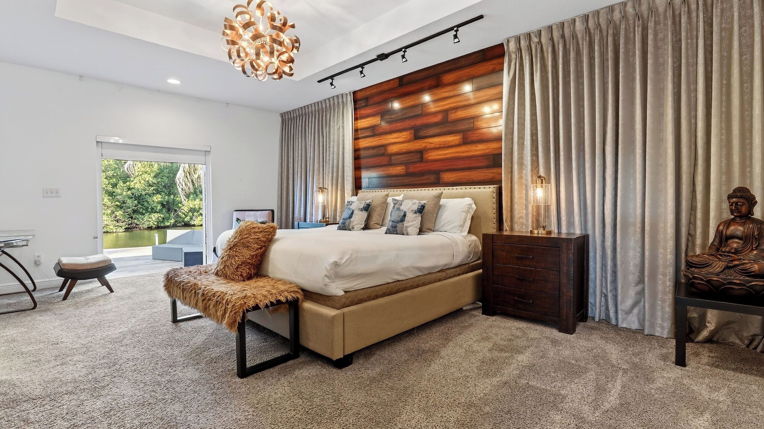 A modern bedroom with a large bed, a wooden accent wall, a view of greenery, and a Buddha statue.