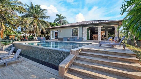 A pool with steps leading to a lounge area, surrounded by palm trees and a building with a stone facade.