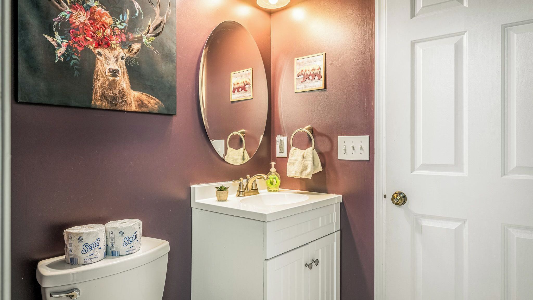 A bathroom with a toilet, sink, and mirror. There are two rolls of toilet paper, a soap dispenser, and a towel hanging on the wall.