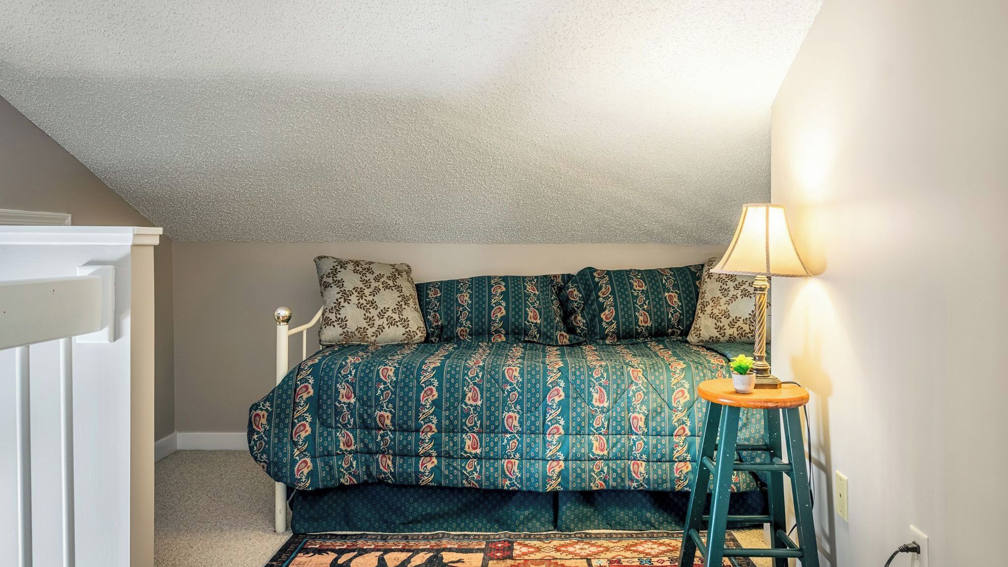 Un dormitorio con cama, mesita de noche, lámpara y alfombra estampada.