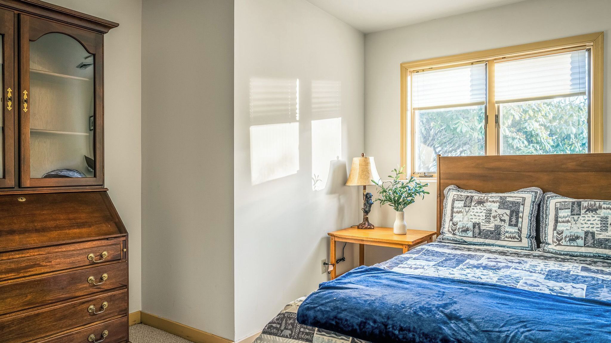 A bedroom with a wooden headboard, a bed with patterned bedding, a nightstand with a lamp, and a dresser with drawers.