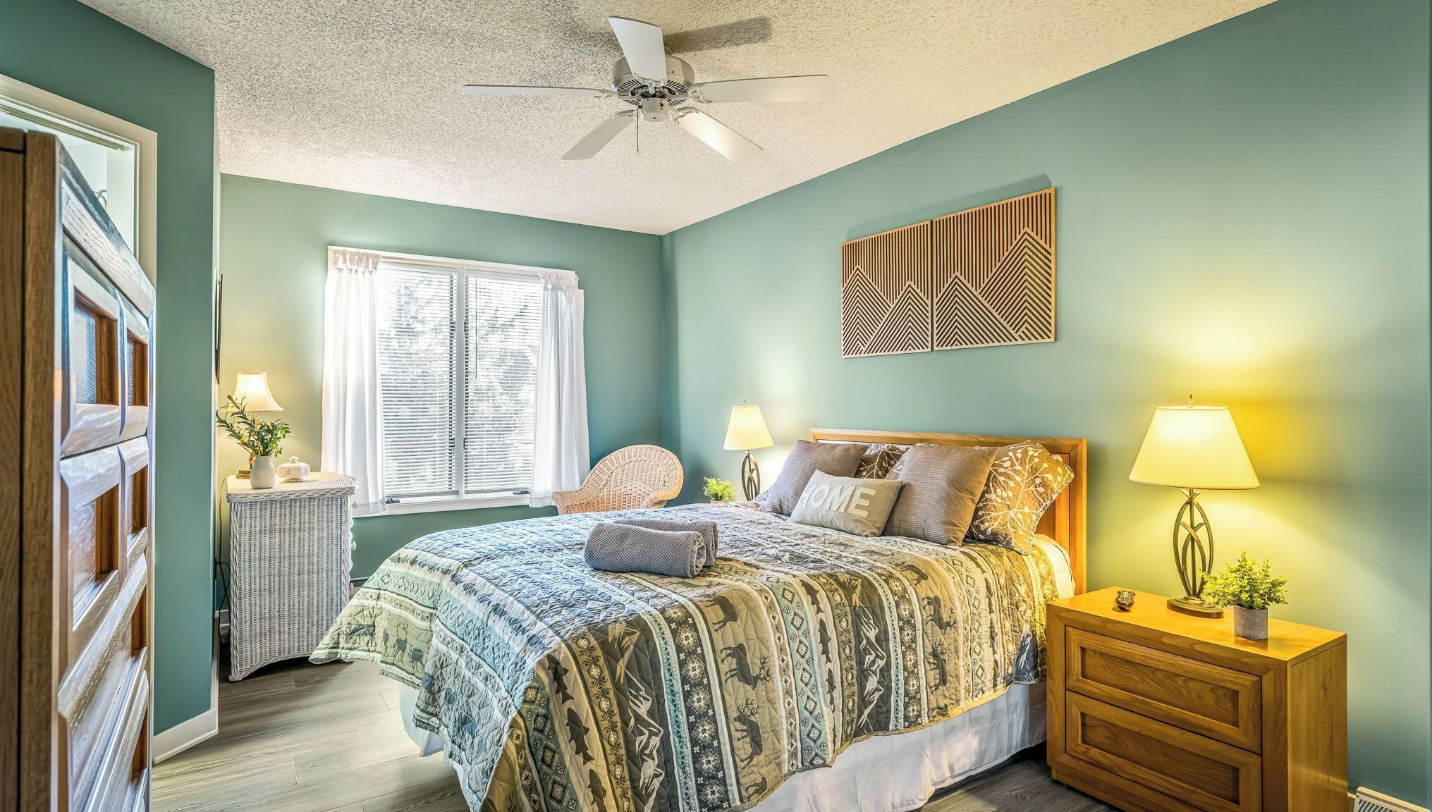A bedroom with a bed, two bedside tables, a chair, a ceiling fan, and a window with blinds.