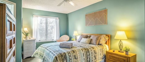 A bedroom with a bed, two bedside tables, a chair, a ceiling fan, and a window with blinds.