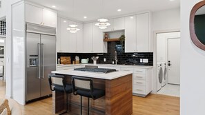 Eine moderne Küche mit weißen Schränken, einem Edelstahl-Kühlschrank, einer Kochinsel mit Barhocker und einer schwarzen Spritzschutzwand.
