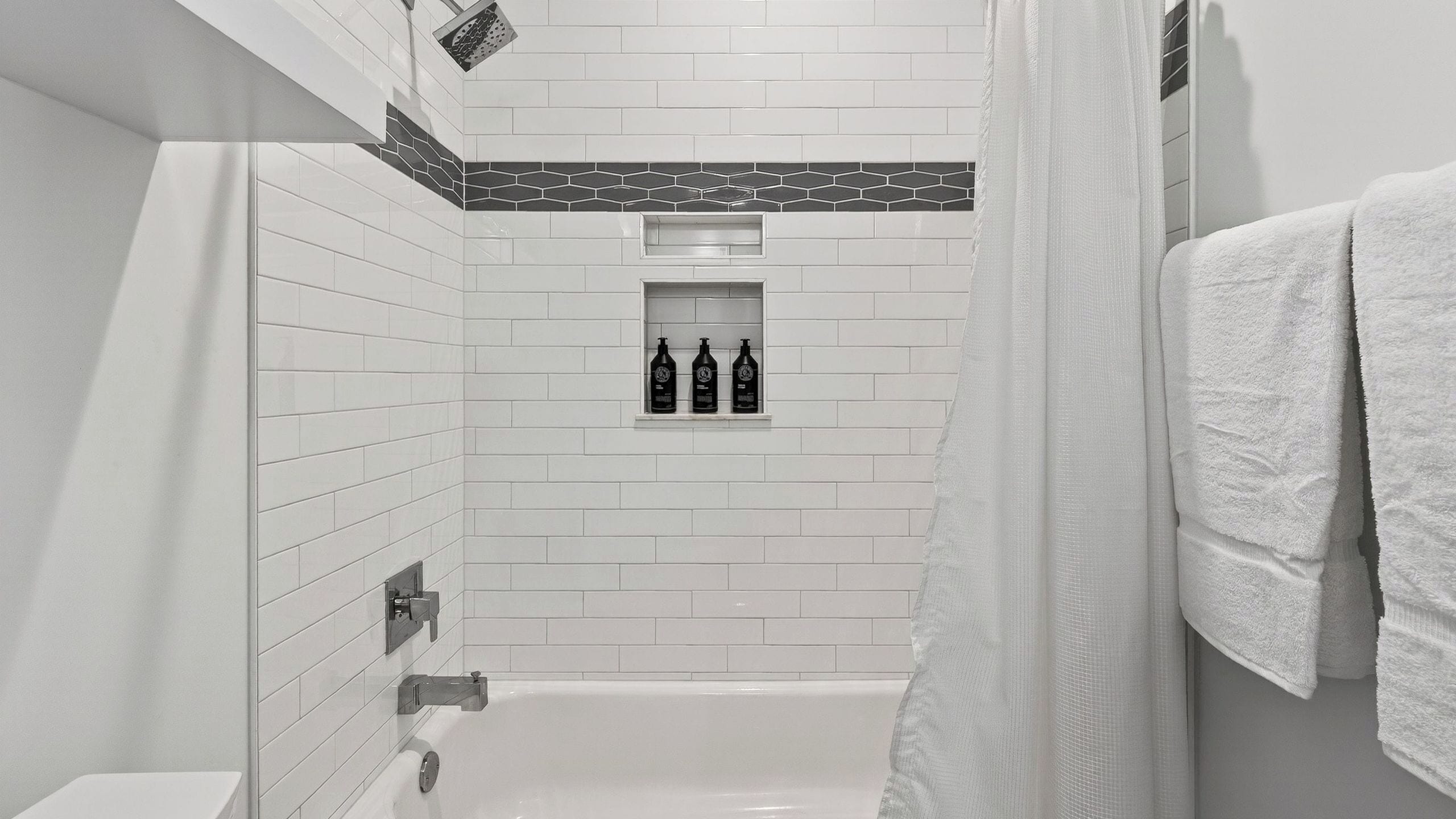 A modern bathroom with white subway tiles, a black and white patterned accent wall, a built-in shelf with three bottles, and a white bathtub.