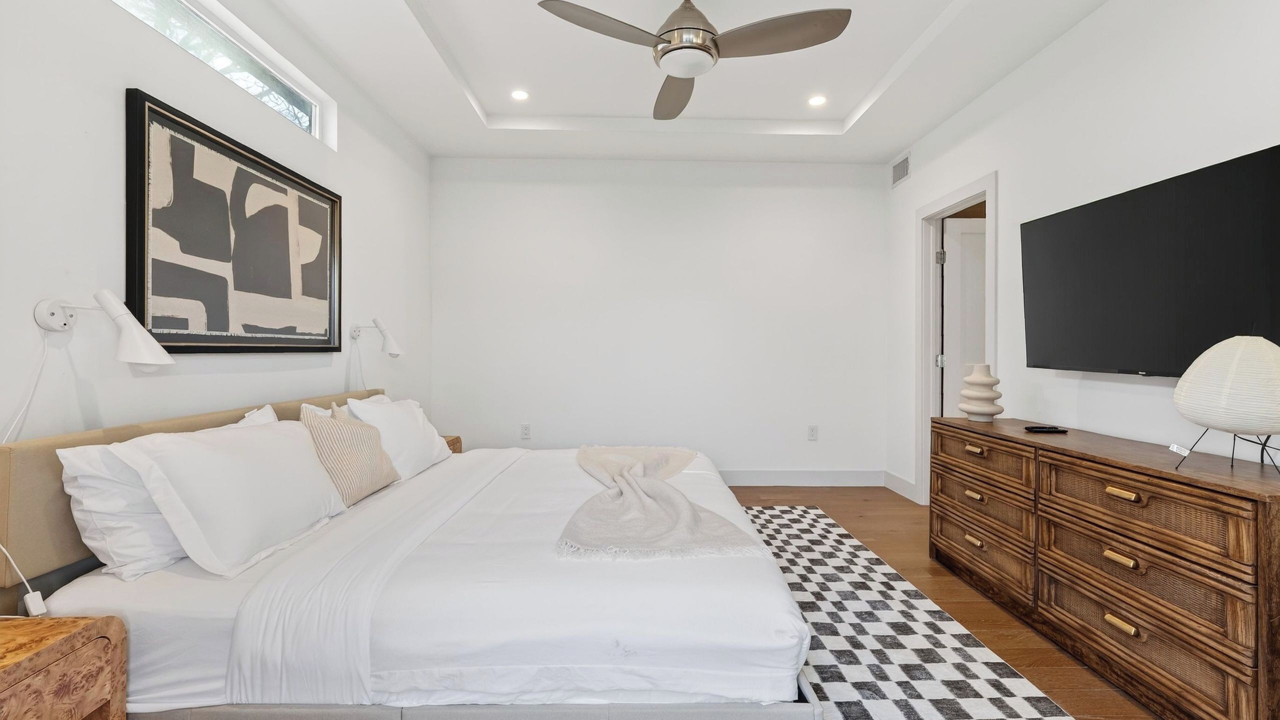 A bedroom with a bed, a ceiling fan, a large TV, and a wooden dresser.