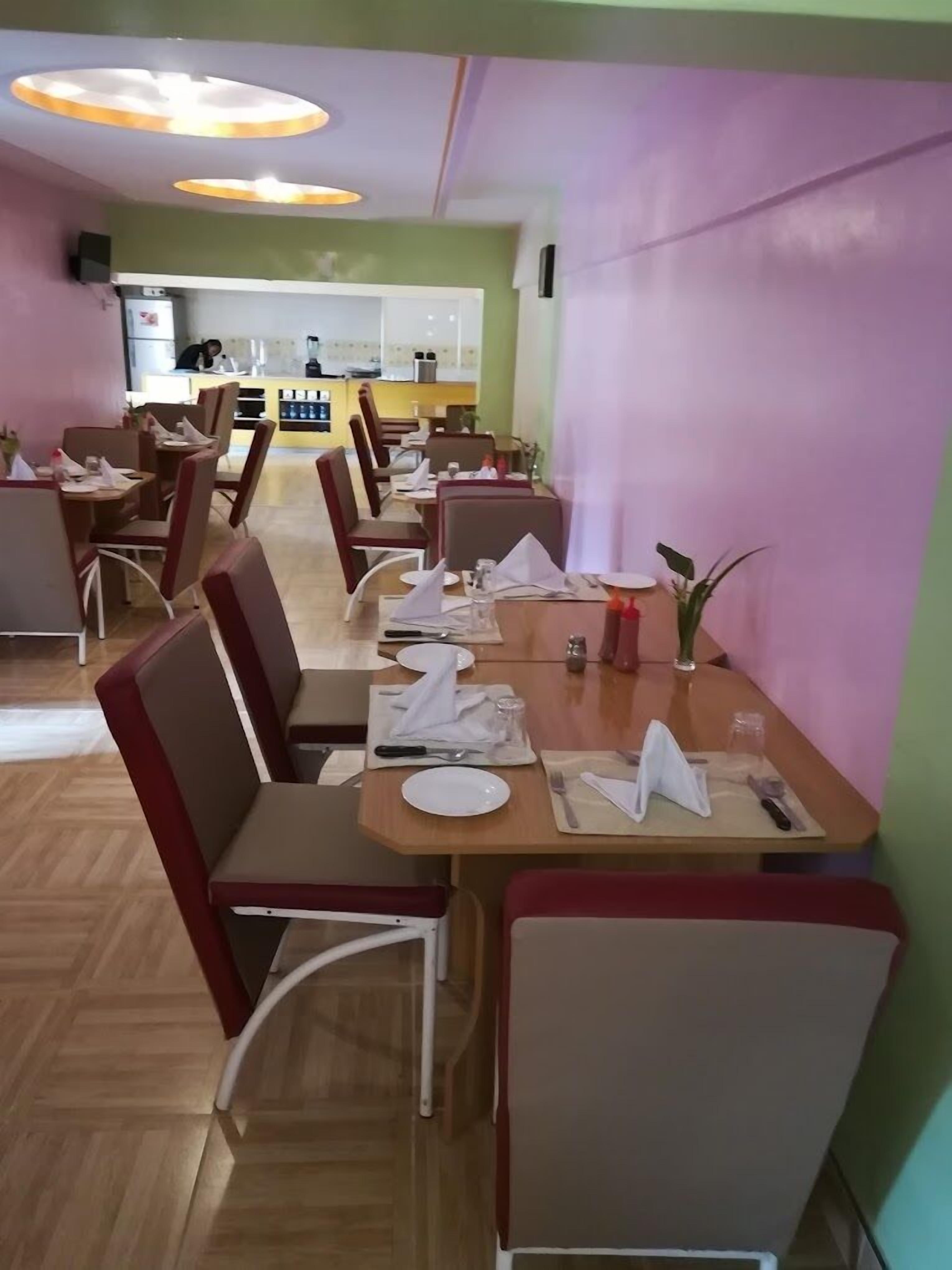Un restaurante con mesas de madera y sillas con cojines rojos, una cocina al fondo y una decoración en tonos verde y morado.