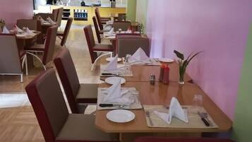 Un restaurante con mesas de madera y sillas con cojines rojos, una cocina al fondo y una decoración en tonos verde y morado.