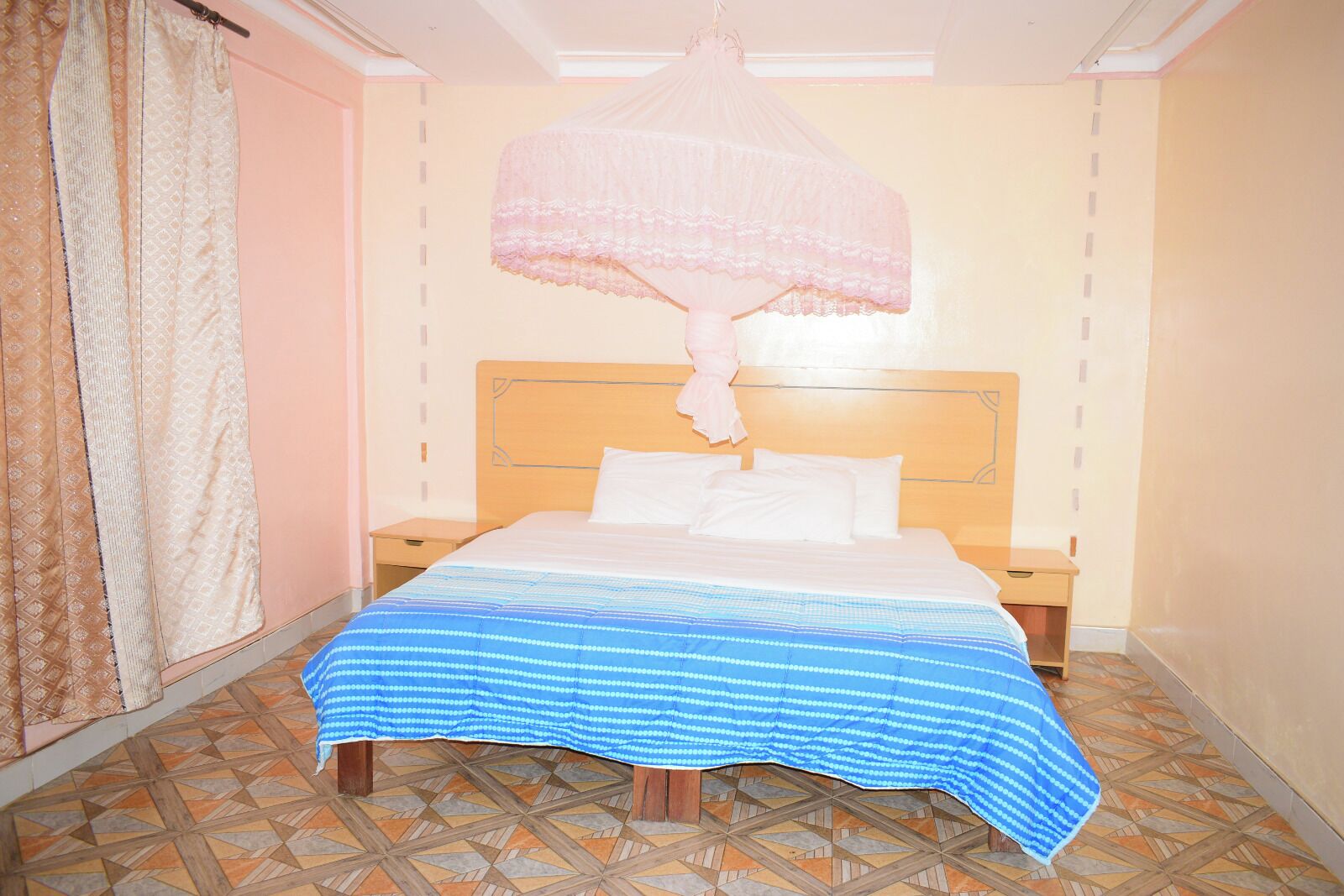 A bedroom with a bed, nightstands, a hanging canopy, patterned curtains, and a tiled floor.