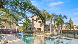 Patio residencial con piscina, palmeras y una casa con jardín cercado.