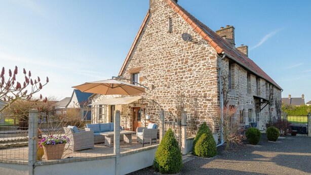 A stone building with a sloped roof, outdoor seating area with a table and chairs, and a small garden with potted plants. - Belle location raffinée (Frênes, Tinchebray-Boc)