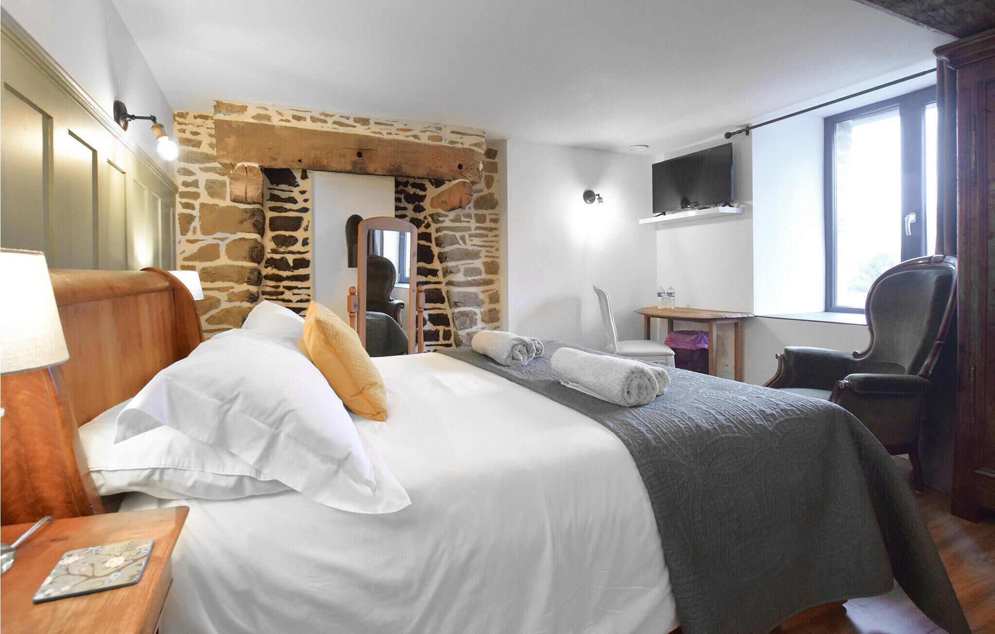 A hotel room with a stone wall, a bed with white linens, a wooden headboard, a desk with a chair, a TV mounted on the wall, and a window with curtains.