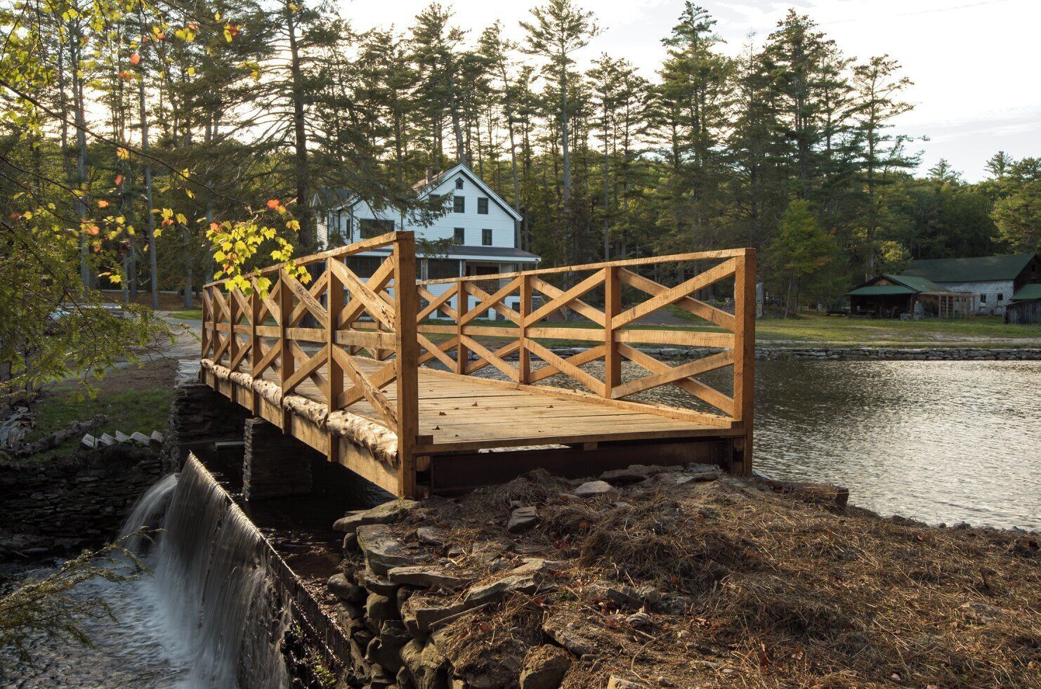 A wooden bridge over a stream leading to a white house with a green roof, surrounded by trees.