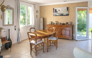 Un coin repas avec une table et des chaises en bois, un meuble contenant de l’ustensile de cuisine et une vue sur la verdure, visible à travers une porte vitrée.