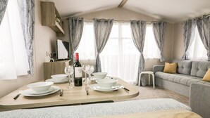 Ein modernes Wohnzimmer mit Essbereich. Es gibt ein Sofa, einen an der Wand montierten Fernseher und einen Esstisch, gedeckt mit Tellern, Gläsern und einer Weinflasche.