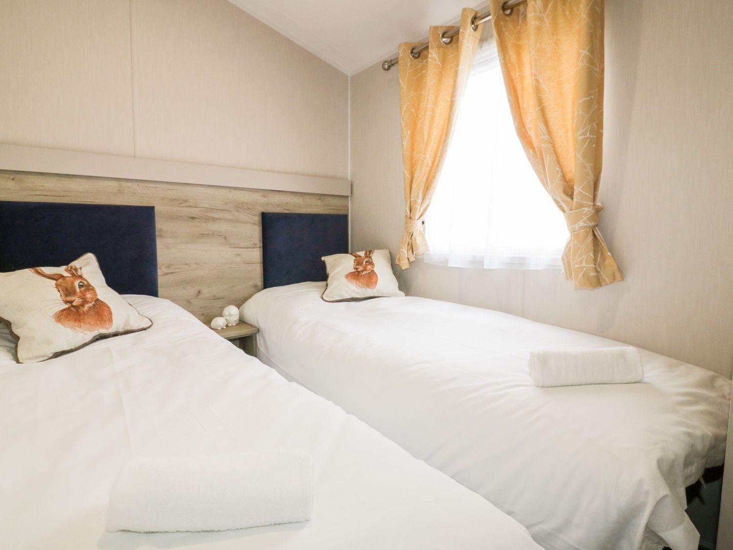 Una camera d'albergo con due letti singoli, ognuno con copripiumino bianco e un cuscino con un motivo a coniglio. La stanza ha una testiera in legno, una finestra con tende gialle e un comodino bianco con una lampada.