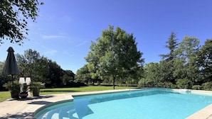 Een rechthoekig zwembad met een helder blauw wateroppervlak, omringd door een verhard terras en een gazon met bomen.