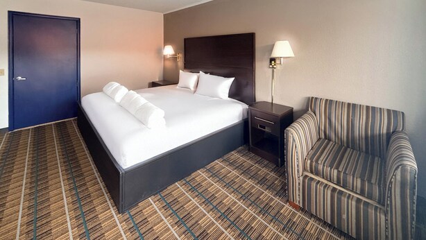 A hotel room with a large bed, two bedside tables, a striped armchair, and a dark-colored door. - Royal INN  (Richmond)