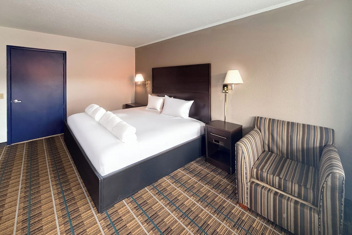 Habitación de hotel con una cama grande, dos mesitas de noche, un sillón a rayas y una puerta de color oscuro.