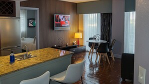 Una habitación de hotel moderna con área de cocina, una mesa de comedor con sillas, un televisor montado en la pared y una ventana con cortinas.
